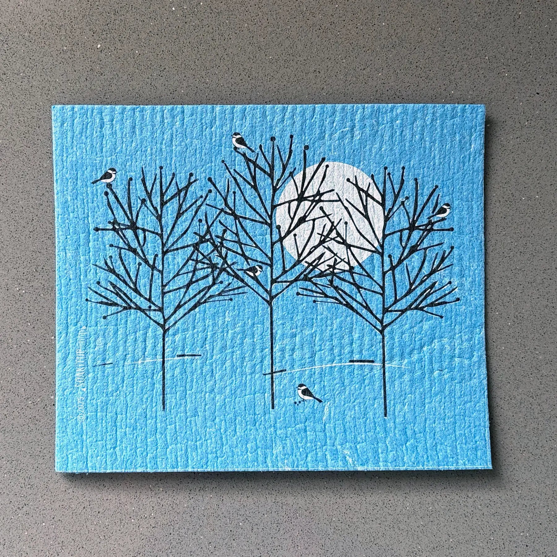 Blue textured reusable dishcloth featuring a minimalist illustration of black-barked trees, birds, and a white moon.