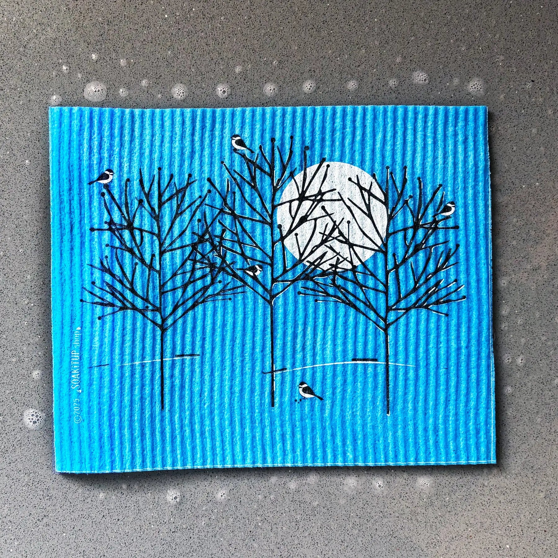 Bright blue corrugated sponge featuring a black-and-white illustration of birds perched on bare trees with a full moon.