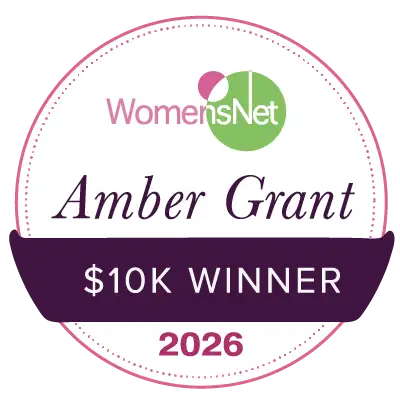A circular, white and purple award badge with a green logo for womensnet, featuring the text ’amber grant’ as ’$10k winner 2026’’.