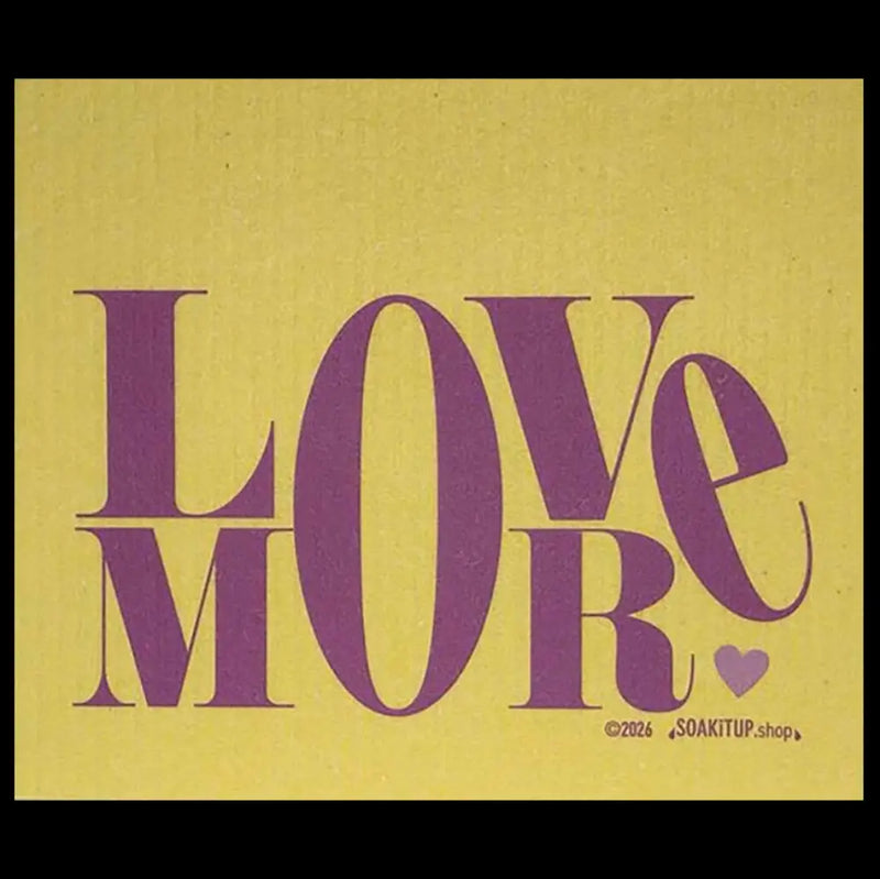 A square Swedish dishcloth featuring the word 'LOVE' in large purple serif letters over a textured mustard-yellow background, with 'MORE' in matching purple below and a small purple heart at the bottom right. Small copyright text reads '©2026 SOAKITUP.