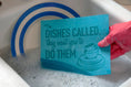 Load image into Gallery viewer, Turquoise swedish dishcloth with humorous dishes called phrase and stacked cup design held in pink glove over soapy water soakitup.shop
