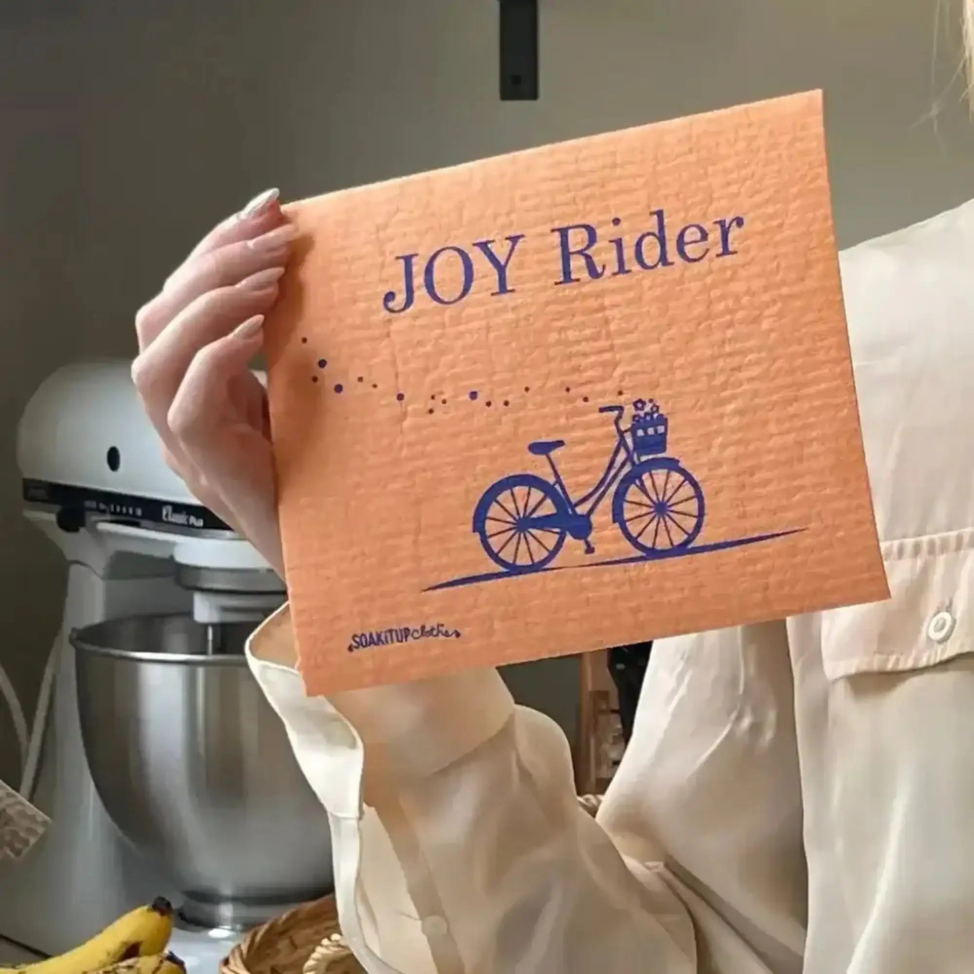 Joy rider bicycle bright orange Swedish dishcloth held by a hand in kitchen setting. Soakitup.shop