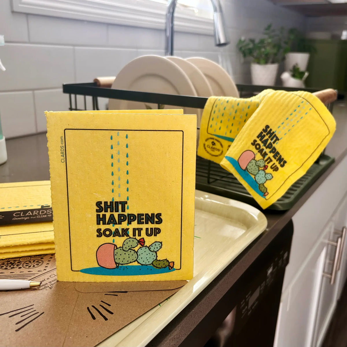 A yellow dishcloth with the text 'SHIT HAPPENS SOAK IT UP' and a cactus illustration stands on a kitchen counter next to other matching dishcloths. Plates and a sink are visible in the background.