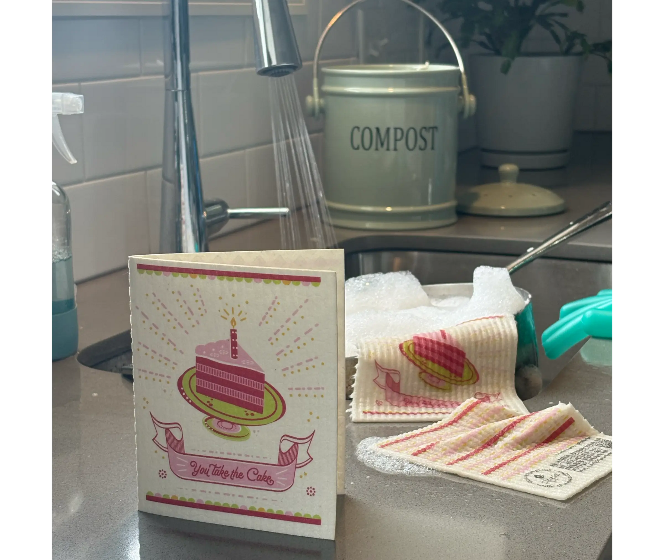 Kitchen counter with a compost bin, cleaning supplies, and a card with a cake illustration. greeting card turns into a cleaning cloth