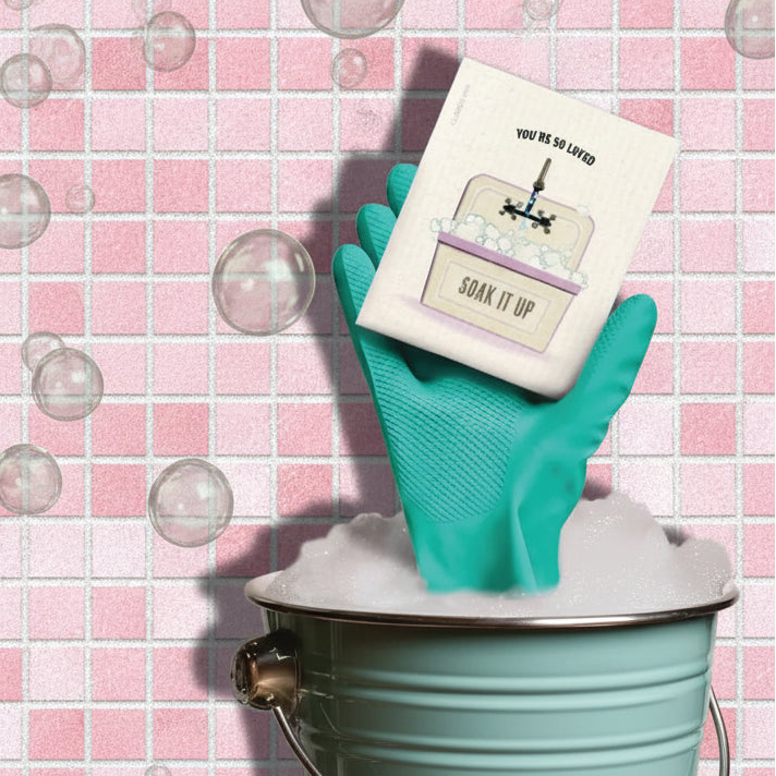 A teal rubber glove holds a white bar of soap labeled ’we’re soaked’ and ’soak it up,’ with an illustration of a bathtub.